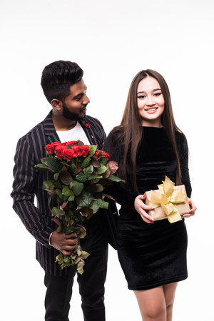 Attractive young asian woman and handsome indian man are standing with gift box and red roses in hands.の写真素材