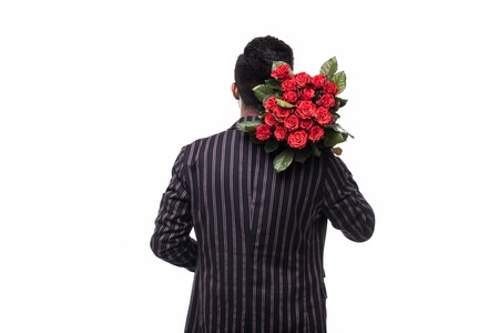 Handsome yound man in suit is standing with red roses behind the back on grey background.の写真素材