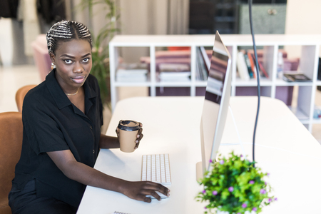 Laughing young African female entrepreneur drinking a cup of coffee and working on a laptop while sitting at a desk in her home officeの写真素材