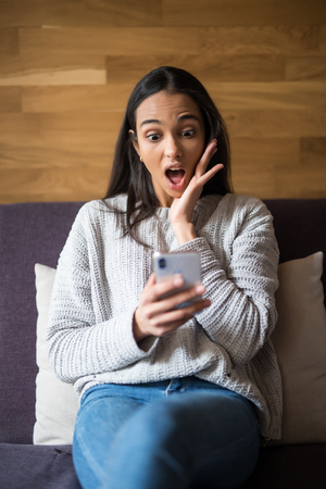 Casual shocked young woman looking at cellphone on sofa at homeの写真素材