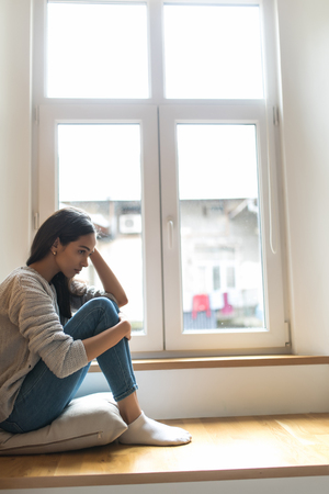 Sad depressed young teenage woman having social problems sitting on windowsillの写真素材