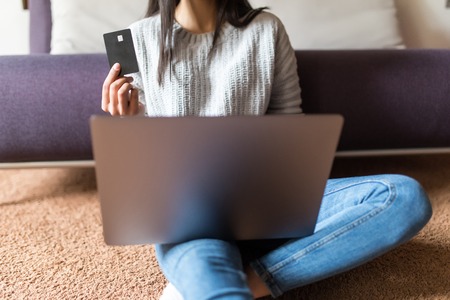 Beautiful young woman working on laptop computer while sitting at the living room, showing credit cardの写真素材
