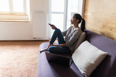 young beautiful woman lying at her apartment living room on sofa couch holding remote control watching television TV conceptの写真素材