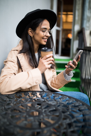 Pretty cheerful young blonde girl in smart casual eclectic outfit sits on cafe terrace while drinking coffee from paper cup. Coffee to goの写真素材