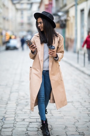 Beautiful smiling woman walking on crowded city street from work with coffee cup and texting on...