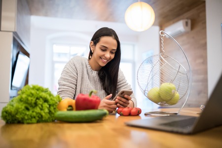 Image of smiling caucasian woman using mobile phone while cooking fresh vegetables salad in kitchenの写真素材