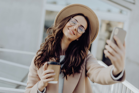 Beautiful young stylish blonde woman wearing a coat walking outdoors, taking a selfie with mobile phone, holding takeaway coffee cupの写真素材