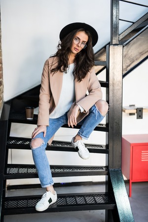 Beautiful thoughtful girl in a coat sitting on the stairs indoorsの写真素材