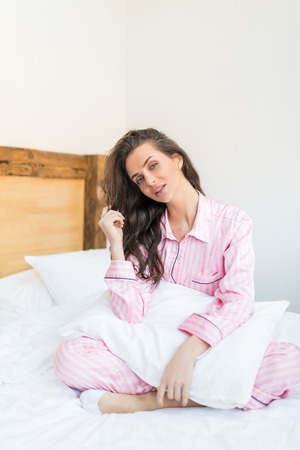 Side view of attractive young smiling woman sitting in bed and hugging a pillowの写真素材