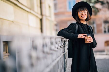 Beautiful young woman holding coffee cup and smiling while walking along the streetの写真素材