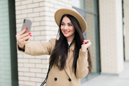 Outdoors lifestyle fashion portrait of pretty young woman making selfie by the phone. Smiling, walking on the city street. Going shopping. Wearing stylish white coat, purple neckscarf, round sunglassの写真素材