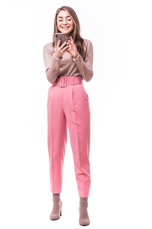 Full length portrait of a young woman typing a text message on her cell phone and leaning against a wall isolated on white background, studio shotの写真素材