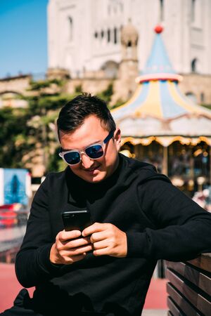 Young handsome man in sunglasses use phone while sitting on the bench.の写真素材