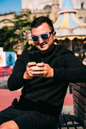 Young handsome man in sunglasses use phone while sitting on the bench.の写真素材
