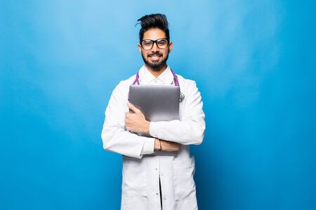 Indian doctor standing in the studio while working with laptop, isolated on blue backgroundの写真素材