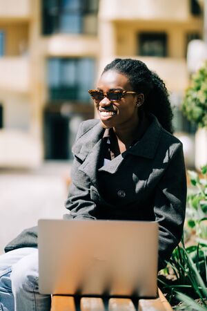 Smiling young African female university student sitting on a campus bench working on a laptop while...