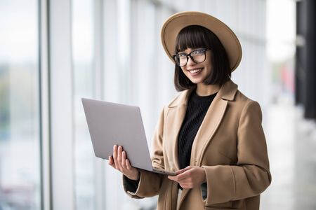 Portrait of woman working holding laptop standing against panoramic window with city view