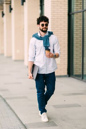 A bearded, smiling, stylish man walking on the streets of the city near modern office centre with laptop in hands and coffee to goの写真素材