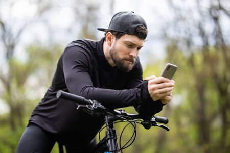 Young sportsman riding bicycle, holding smartphone, sunny autumn parkの写真素材