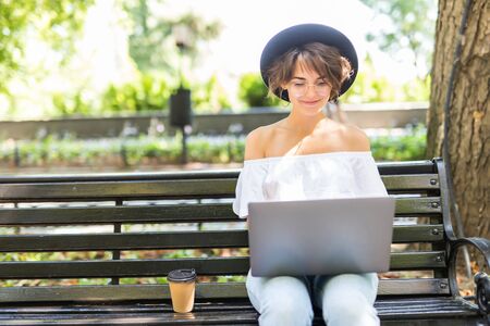 Young student with a tablet and a disposable coffee cup sitting on the bench and reading in a summer park.の写真素材