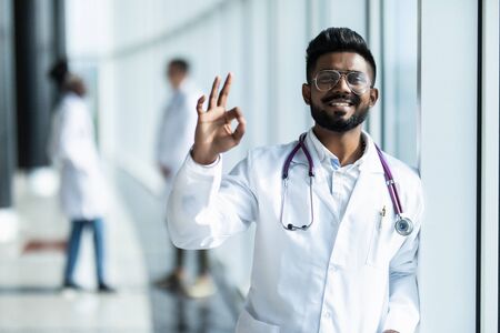 Young indian male doctor with okay sign in white uniform with colleguesの写真素材
