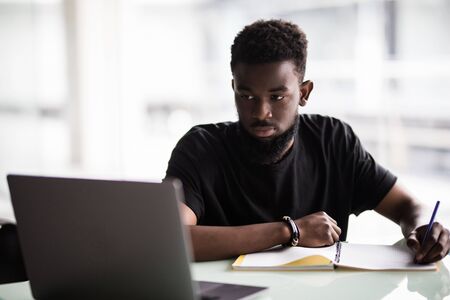 Image of young African man looking at camera with laptop near by