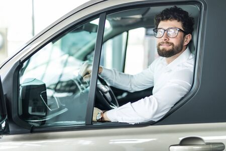 Handsome bearded mature man sitting relaxed in his newly bought car looking out the window smiling...