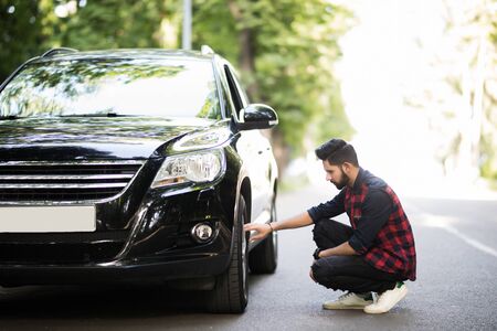 Cheerful young businessman has problems with the wheel of his car. He is kneeing and looking at it...