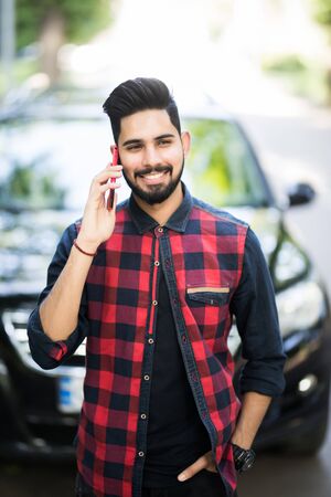 Portrait of indian man wearing suit, standing near black car and talking on smartphone