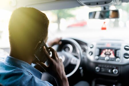 Handsome cheerful African man talking on his smart phone, sitting in a car on a countryside road.の写真素材