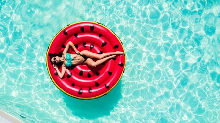 Aerial top of woman wearing a hat sitting in inflatable watermelon in deep cyan calm waters like a...