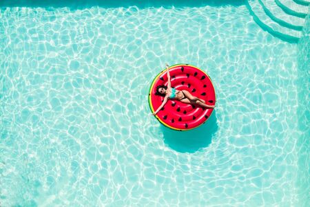 Aerial top of woman wearing a hat sitting in inflatable watermelon in deep cyan calm waters like a...