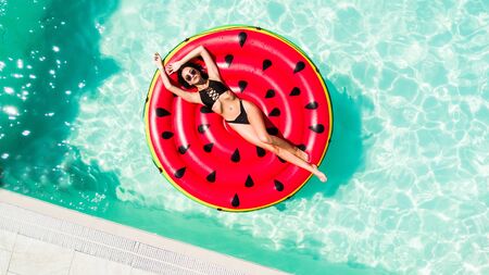 Aerial view of female in bikini lying on a floating inflatable mattress watermelon in swimming pool...