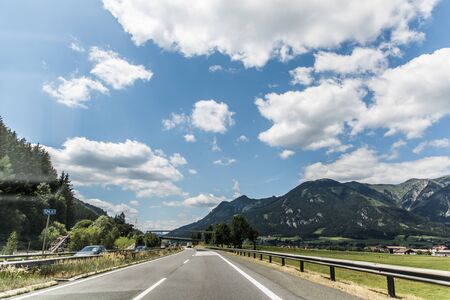 Autobahn or highway with a bridge in the mountains with clear marking surrounded by vibrant green trees under blue sky.の写真素材