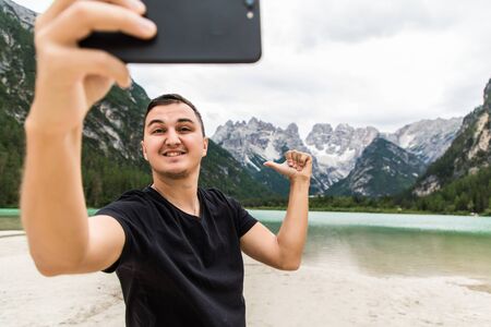 Young man in high mountain near alpine lake taking selfie portrait with beautiful landscape.
