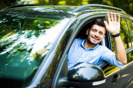 Portrait of young handsome brunette man driving car and greeting somebody with hand.