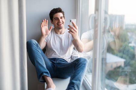 Young man sitting on the windowsill make video call on the phoneの写真素材