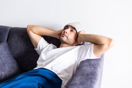 Sick man lying on sofa checking his temperature under a blanket at home in the living roomの写真素材