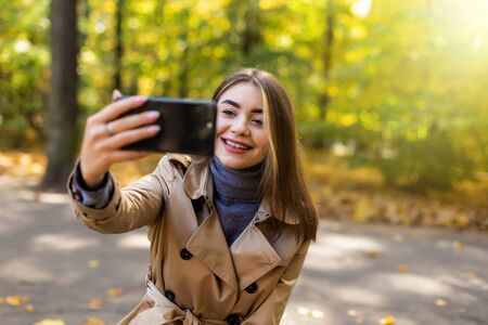 Positive woman making selfie while drinking coffee in autumn park.
