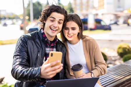 Young couple working on laptop and make video call on phone smiling while sitting relaxed on bench...