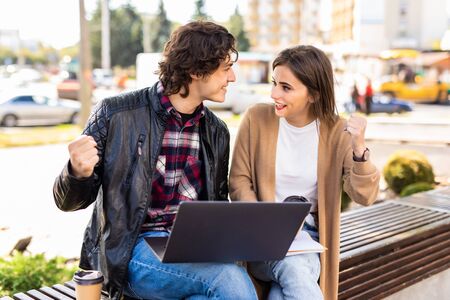 Happy young couple using laptop computer sitting on a bench in city outdoor - Two lovers having fun spending time together to watch social network - Concept of love, people, addiction, technologyの写真素材