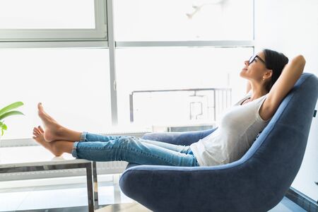 Young woman at home sitting on modern chair in front of window relaxing in her living roomの写真素材