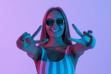 Young woman pointed peace gesture wearing striped pink swimsuit and sunglasses over pink background