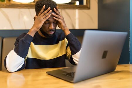 Young african man looking at laptop, thinking, decided problem, have a stress at cafe. Freelancer job.の写真素材