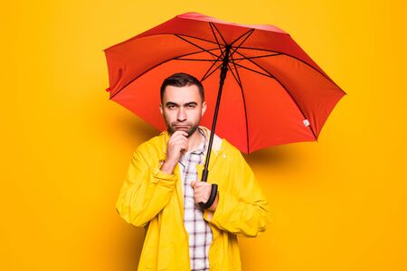 Young handsome bearded man in yellow raincoat with red umbrella thinking over orange backgroundの写真素材