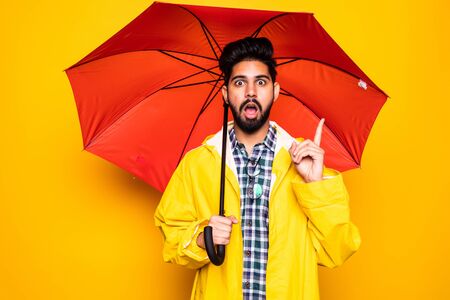 Young handsome bearded indian man in yellow raincoat with red umbrella cover from rain have idea over orange backgroundの写真素材