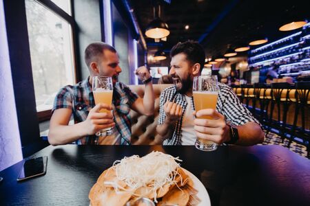 Two cheerful excited young men watching match on laptop and supporting their team sitting in barの写真素材