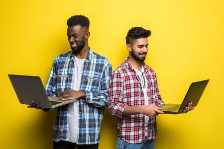 Portrait of a two happy young men holding laptop computer while standing and celebrating isolated over yellow backgroundの写真素材