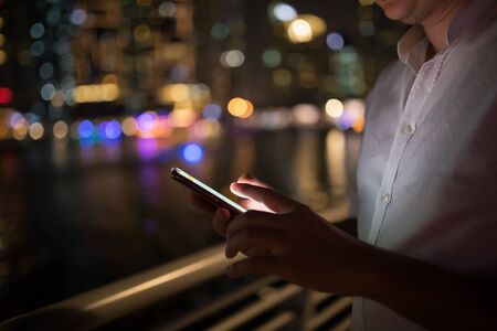 Close-up image of male hands using smartphone at night on city shopping street. Searching or social...