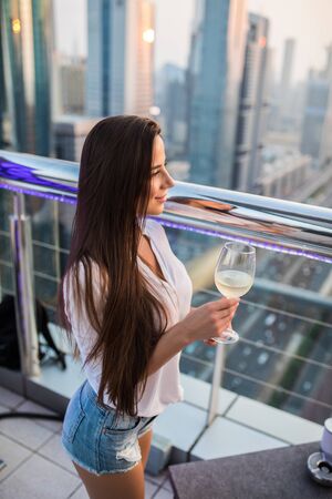 Young woman with glass of wine while enjoying the view at the top at skybar on skyscrapers and sunsetの写真素材